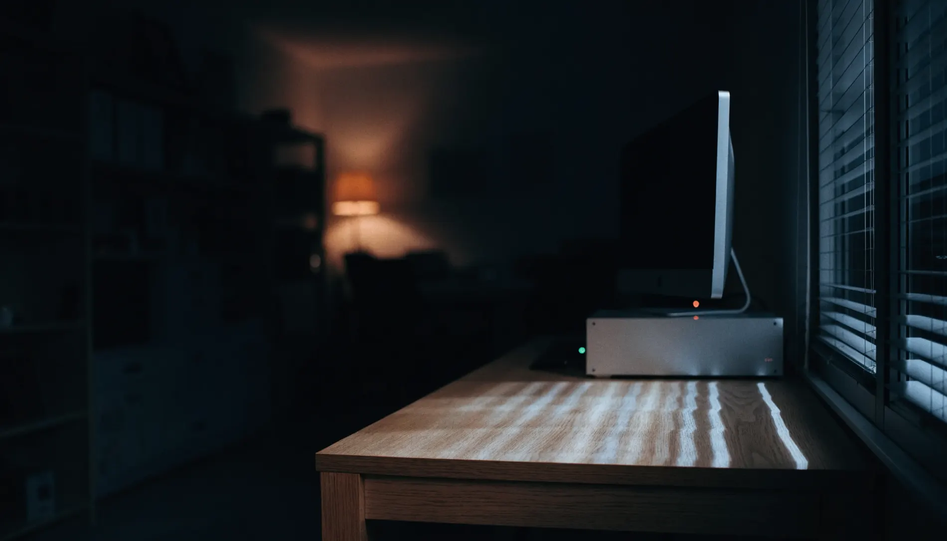A dark, empty home office at night. A desktop machine on an oak desk sits awake, its status light on, with nobody in the room.